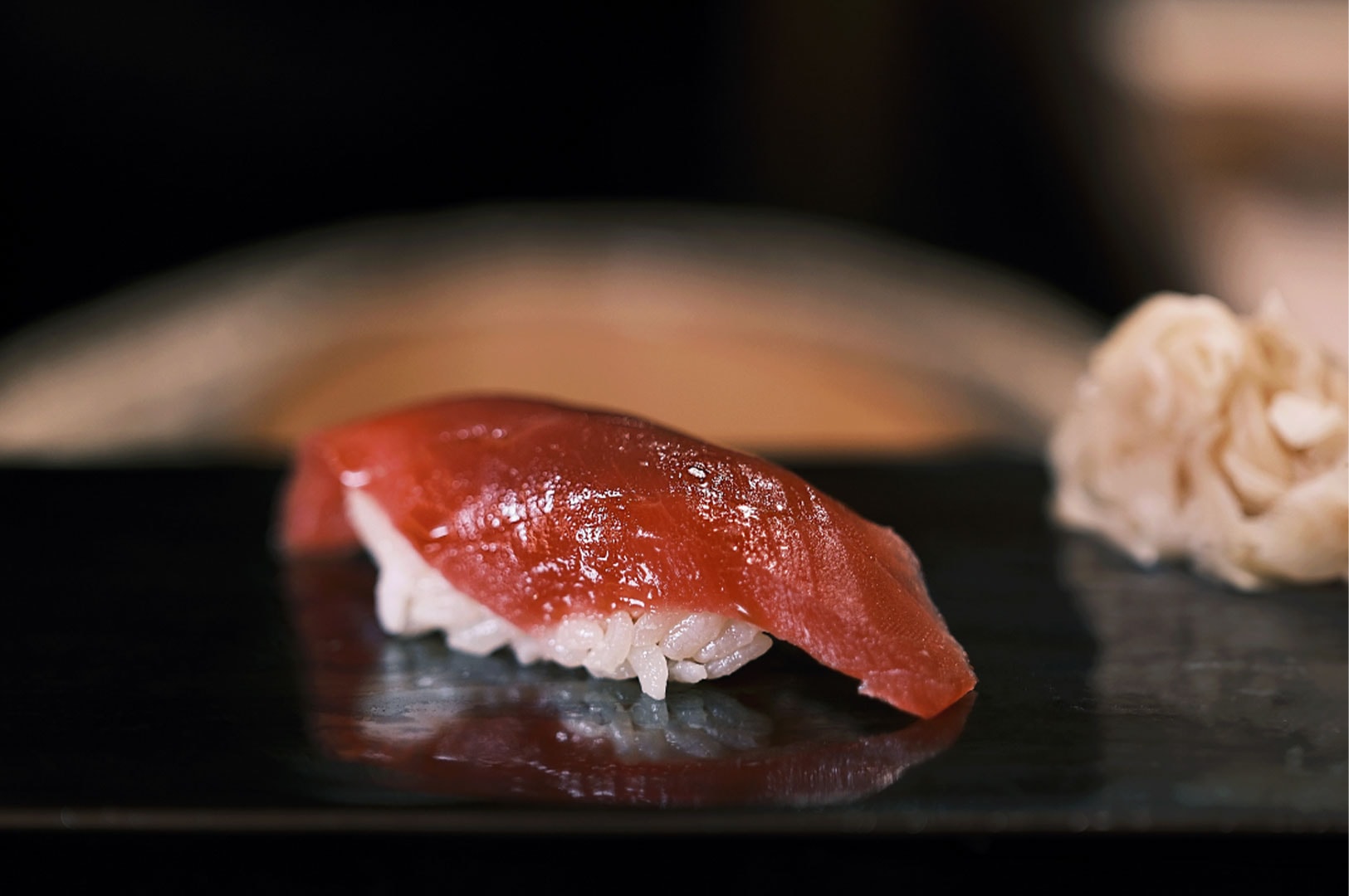A single piece of glistening red tuna (maguro) nigiri sushi served on a black plate with pickled ginger in the background.