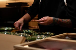 A chef wearing a dark traditional uniform meticulously prepares multiple dishes, with their hands focused on placing delicate yellow garnishes into a row of ornate green and gold ceramic bowls. The chef’s left forearm is adorned with a vibrant, detailed tattoo featuring traditional Japanese floral and wave motifs. In the foreground, wooden boxes containing fresh ingredients are partially visible on the clean, light-colored wooden counter.