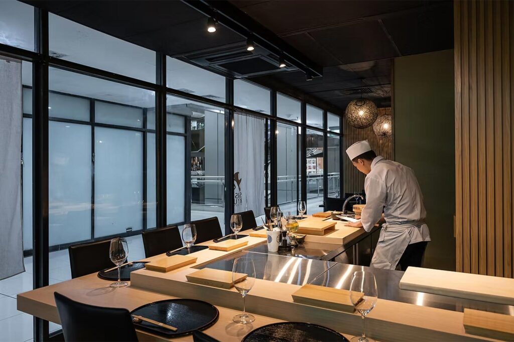 A chef in a traditional white uniform working at a minimalist light-wood counter in a restaurant with large glass partitions.