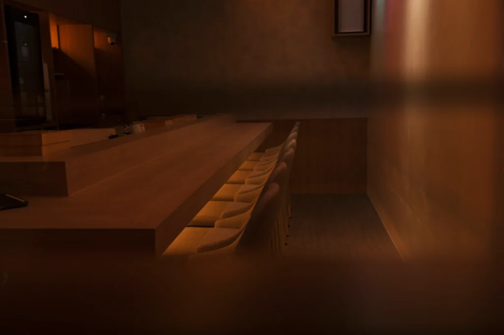 A long wooden bar with several chairs, illuminated softly in a dark setting of a Real Sushi Omakase restaurant.