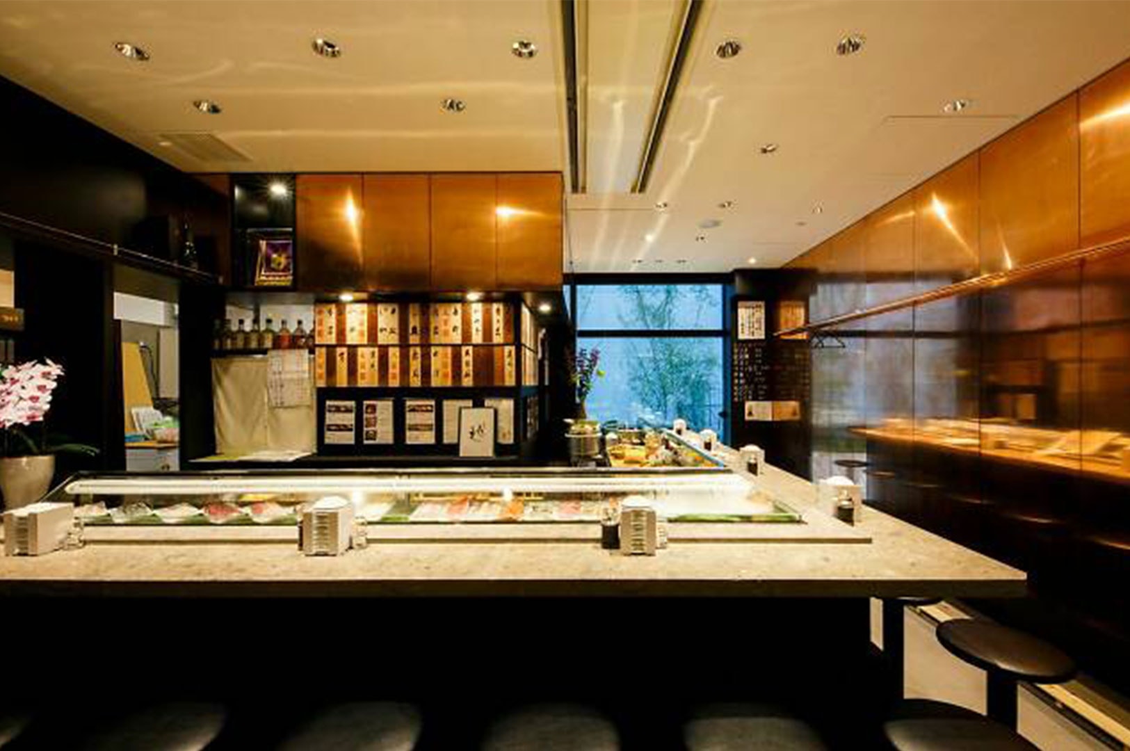 A wide view of an inviting sushi bar counter featuring warm lighting, copper wall accents, and wooden menu plaques hanging on the back wall.