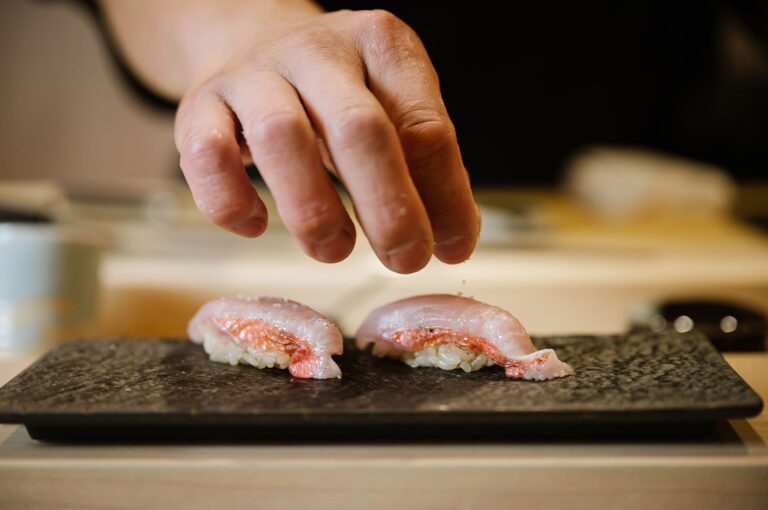 A close-up of a sushi chef's hand sprinkling coarse salt seasoning onto two pieces of fresh nigiri served on a textured black plate.