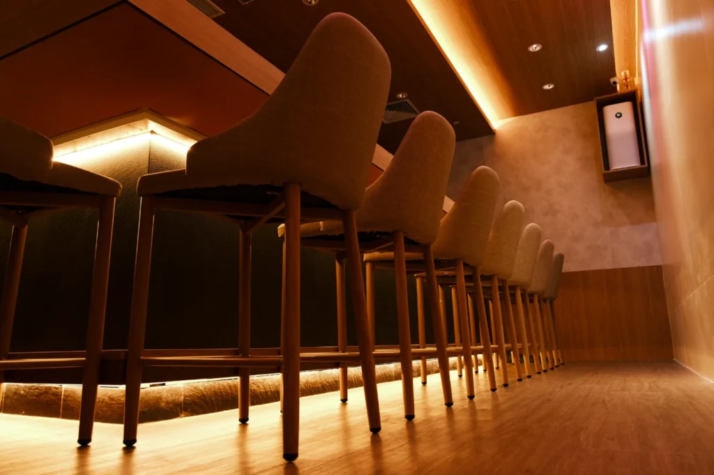 A low-angle shot captures a row of elegant, high-backed bar stools with light gray upholstery lined up against a dark sushi counter. Warm, ambient cove lighting glows from beneath the counter and along the ceiling, highlighting the polished wooden floors and minimalist interior design. The perspective creates a sense of depth, leading the eye down the long, inviting seating area of the omakase restaurant.