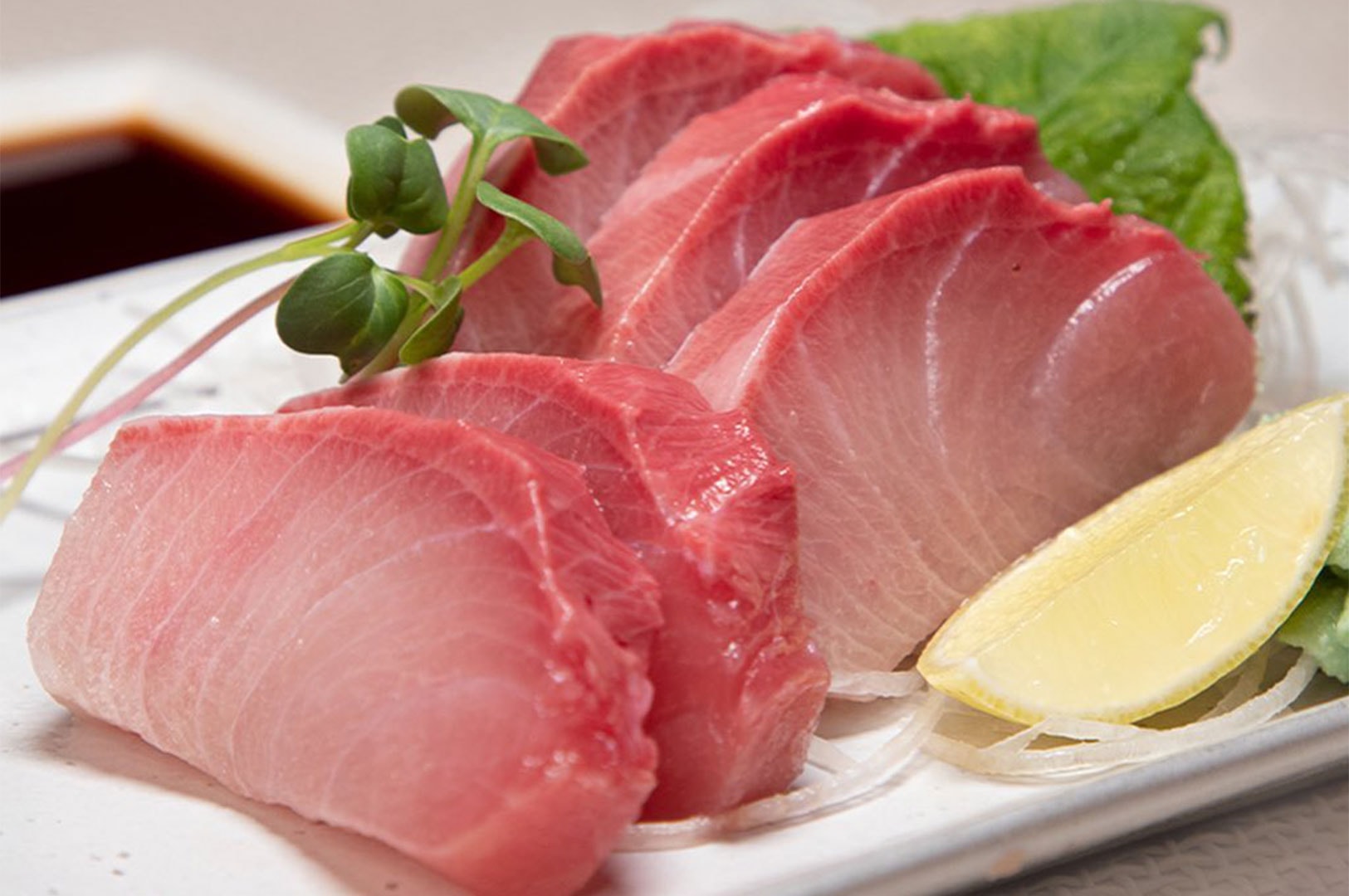 A close-up of thick, fresh slices of pink Yellowtail sashimi garnished with green sprouts and a lemon wedge.