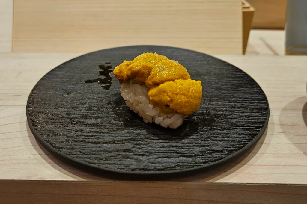 A single piece of Uni (sea urchin) nigiri sushi is presented on a textured, circular black stone plate. The bright orange, creamy lobes of sea urchin are generously piled atop a small bed of seasoned white sushi rice. The plate sits on a light-colored wooden sushi counter, emphasizing the simple, high-quality craftsmanship of the dish.