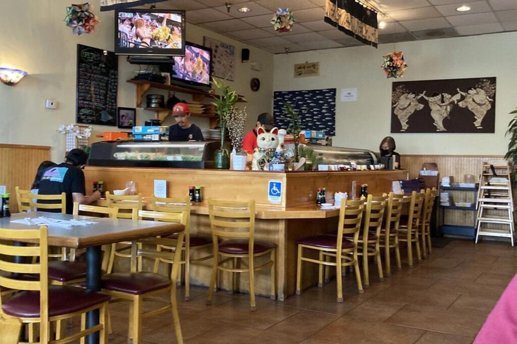 Interior of a casual, traditional sushi restaurant featuring a wooden L-shaped counter, Maneki-neko lucky cat statue, and wall-mounted TVs.