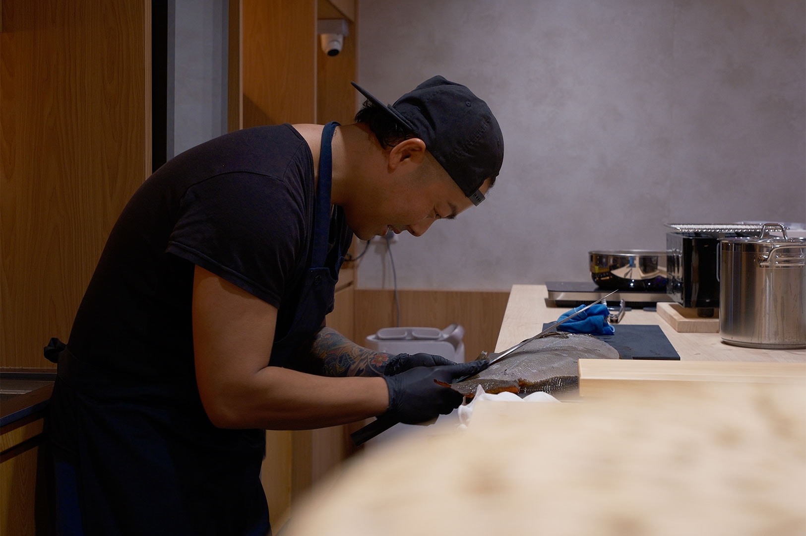 A sushi chef wearing black gloves using a long knife to carefully fillet a large flatfish (flounder/hirame) for sashimi preparation.
