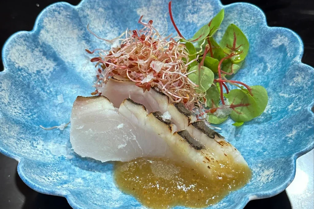 A beautiful blue, flower-shaped ceramic bowl holds several slices of seared white fish sashimi. The fish is accompanied by a mound of pinkish micro-sprouts, small green leaves, and a dollop of translucent, savory citrus-based sauce at the base. Fine flakes of sea salt are scattered over the fish, highlighting the fresh, artistic presentation of the course.