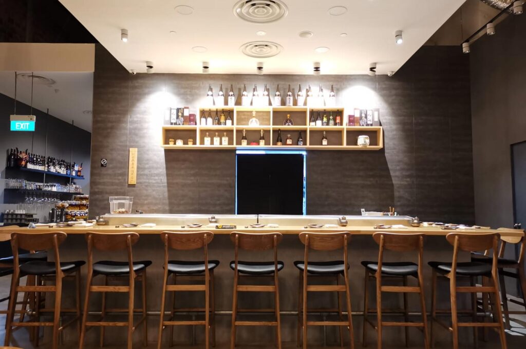 Symmetric view of an upscale sushi bar with a light wood counter, high-back stools, and a back-lit display shelf of premium sake bottles.