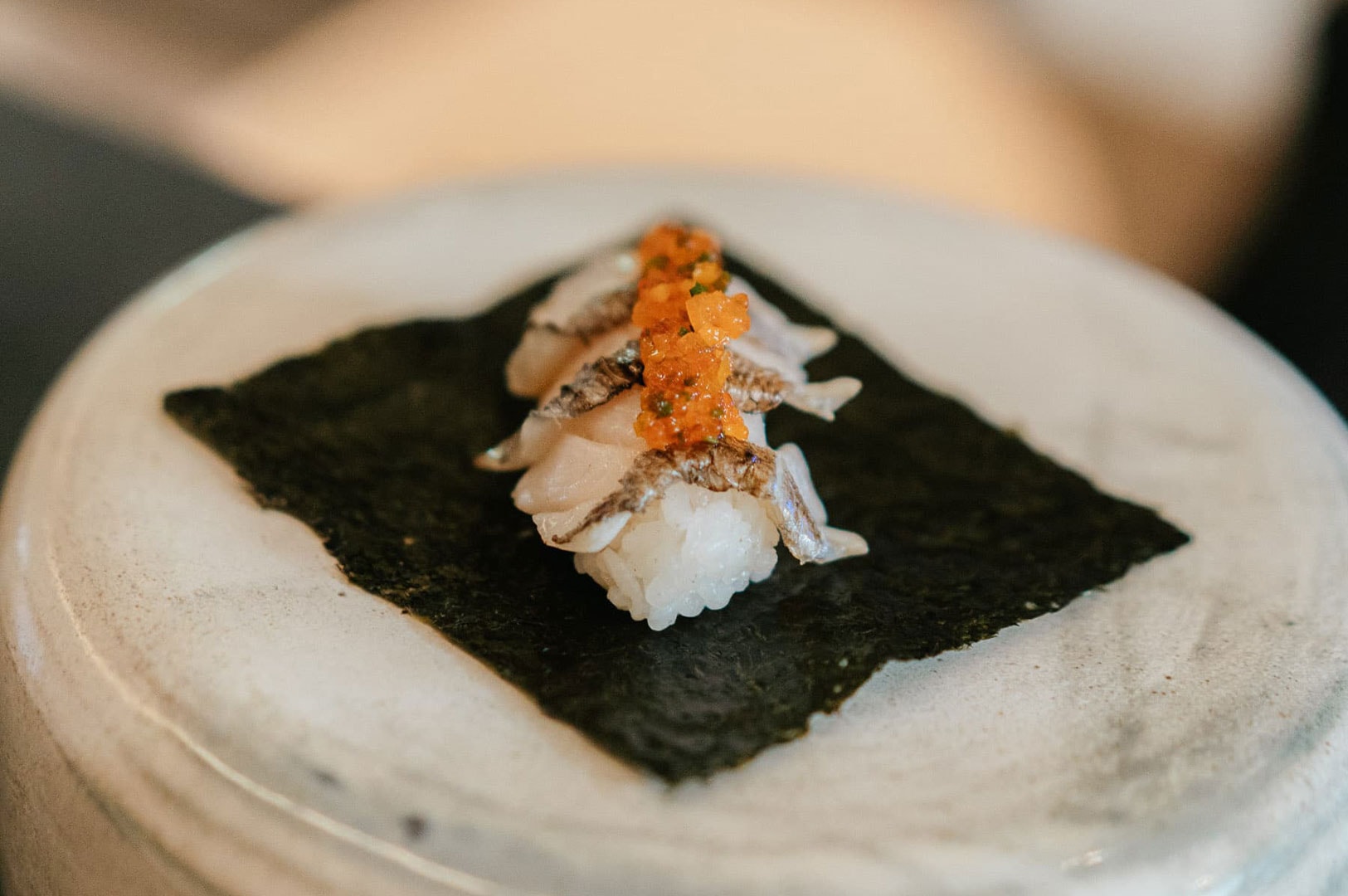 An artistically plated piece of white fish sushi served atop a square sheet of nori seaweed, topped with bright orange garnish.