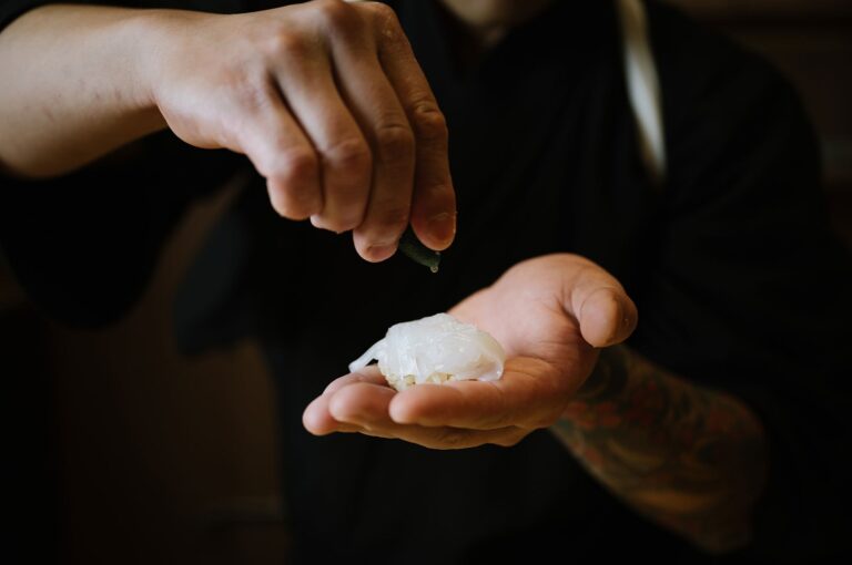 A close-up of a sushi chef's hands squeezing fresh citrus juice over a piece of white fish nigiri topped with seasoning.