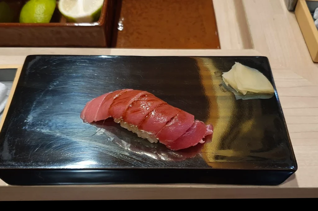 A piece of Maguro (tuna) nigiri sushi, featuring a deep red, glossy slice of fish with delicate score marks, is placed on a rectangular black lacquered platter. A small serving of pale, sliced pickled ginger sits in the corner of the plate. The background shows a clean wooden counter environment with a glimpse of fresh limes, indicating a professional sushi preparation station.
