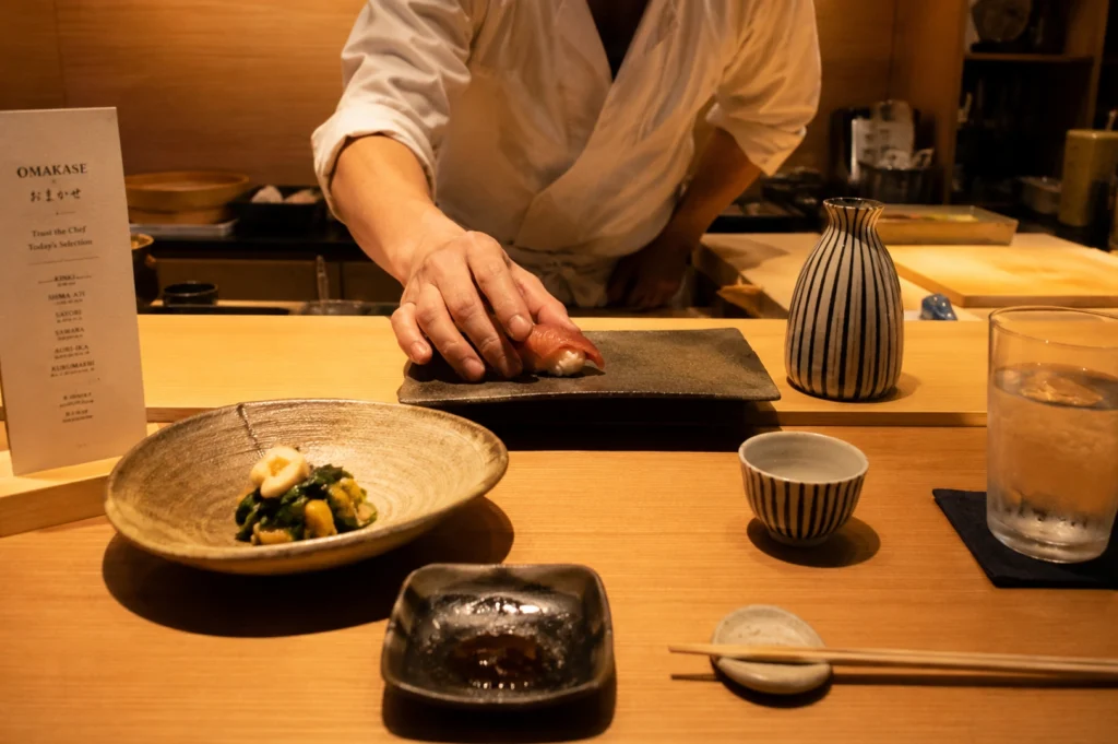 An eye-level, medium shot captures a chef in a white uniform assembling a piece of sushi at a wooden counter. The chef's hands are focused on placing a piece of nigiri with a translucent pink topping onto a dark, rectangular plate. To the left, a small, rustic ceramic bowl holds a green vegetable and corn dish, while a menu card stands upright nearby. On the right, a matching striped ceramic sake flask and small cup sit near a glass of iced water. In the foreground, a small dish with soy sauce and a pair of wooden chopsticks resting on a small holder complete the traditional Japanese dining setting. The warm lighting enhances the rich wood tones of the counter and background.