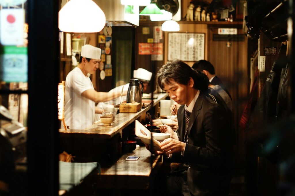 Gemini said A candid, eye-level photograph captures the warm, intimate atmosphere of a narrow Japanese ramen shop or izakaya at night. In the foreground, a man in a dark business suit sits at a weathered wooden counter, his head bowed as he intently reads a menu. Behind the counter, a chef wearing a traditional white uniform and tall toque is captured in motion, preparing food under the glow of low-hanging pendant lamps. The background is filled with cozy, cluttered details, including handwritten menus on the walls, a thermal carafe, and small condiment containers, while the soft, amber lighting creates a moody, cinematic contrast against the dark shadows of the restaurant's interior.