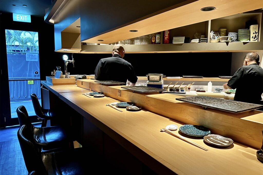 View from behind a light wood omakase counter showing two sushi chefs in black uniforms preparing food under warm pendant lighting.