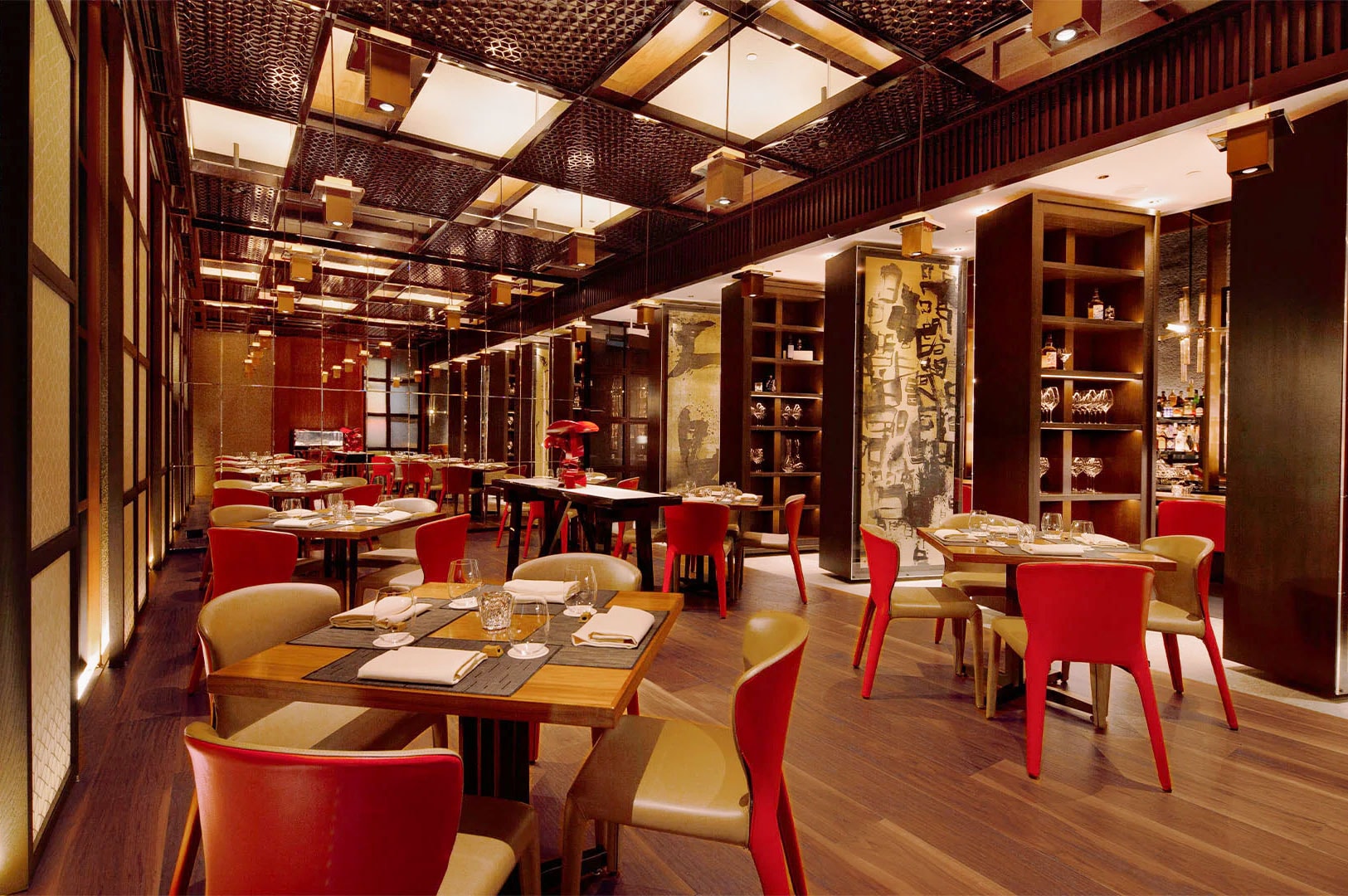 The interior of an upscale restaurant featuring modern wooden tables, red chairs, intricate ceiling latticework, and ambient lighting.
