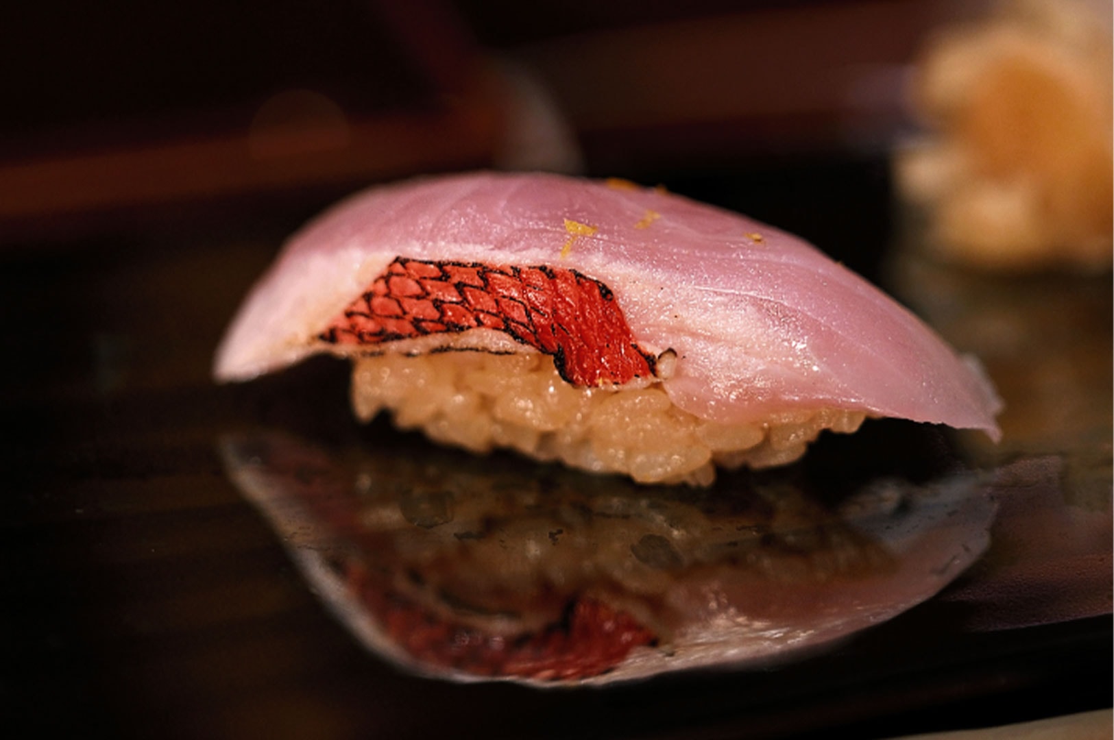 Close-up macro shot of premium Kinmedai (Golden Eye Snapper) nigiri sushi with seared skin and glaze on a reflective black surface.