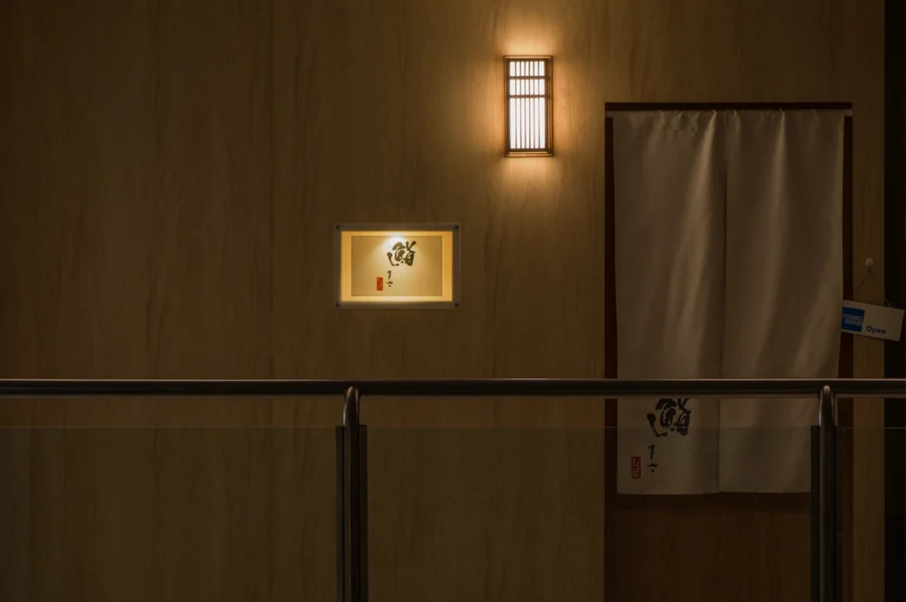 The entrance of a high-end Japanese restaurant features a minimalist wooden wall with a warm, backlit rectangular sign displaying Japanese calligraphy. To the right, a traditional white fabric noren curtain hangs over a doorway, while a small lantern-style wall light above casts a soft glow. A sleek metal and glass railing is visible in the foreground, suggesting an elevated walkway or mall location.