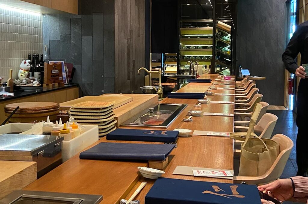 Long perspective of a polished wood sushi counter set for service with blue menus, small ceramic dishes, and a stone wall backdrop.