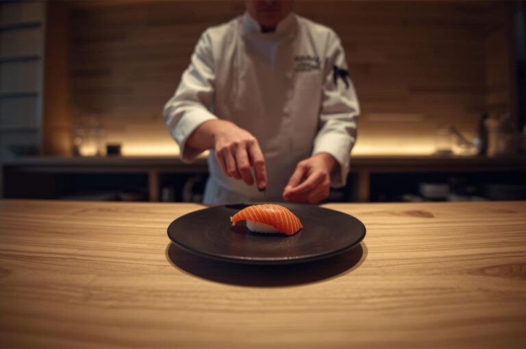 A sushi chef in a white uniform carefully placing a garnish on a piece of fresh salmon nigiri served on a black plate.