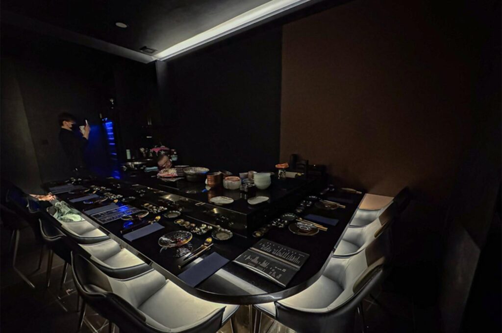 High-contrast, moody dining room with a black U-shaped omakase counter, silver modern chairs, and low-key atmospheric lighting.