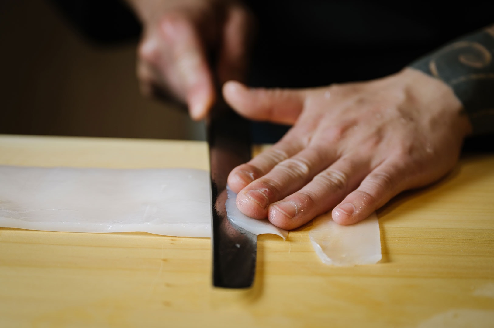 A sushi chef using a traditional knife to precisely slice a fresh fillet of white squid (ika) on a wooden cutting board.