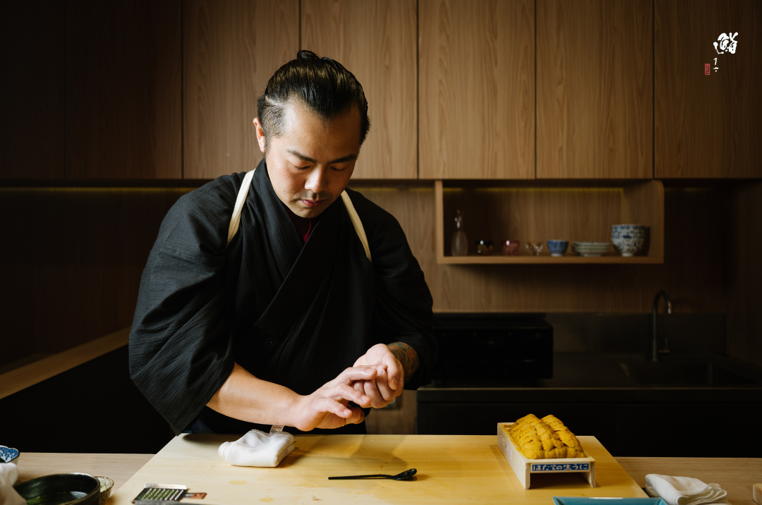 A chef in black attire prepares ingredients on a wooden cutting board at Sushi Masa, highlighting true Japanese culinary art.