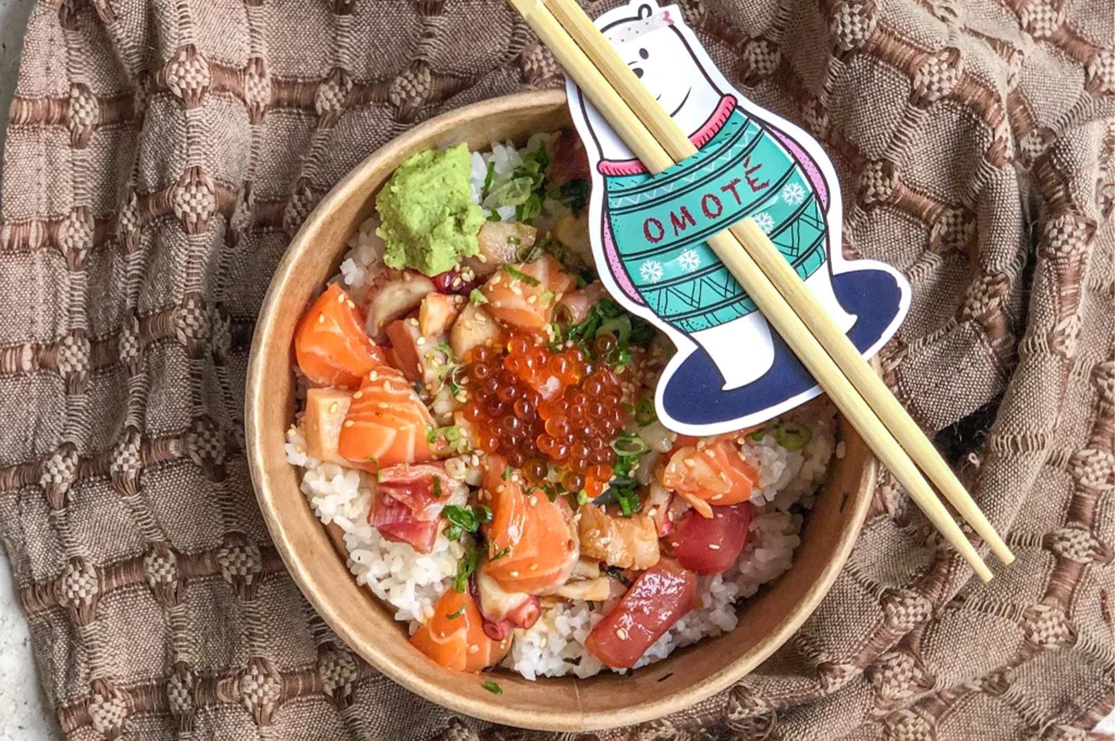 An overhead shot of Omote's chirashi don in a to-go bowl, showcasing mixed raw fish, ikura, and chopsticks held by a festive polar bear mascot.
