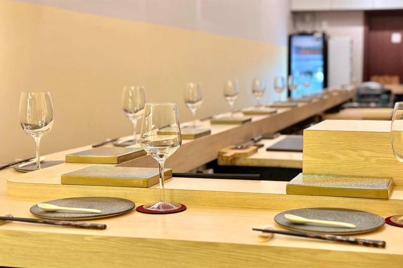 An elegant, light-wood omakase sushi bar counter featuring minimalist place settings with wine glasses, ceramic plates, and chopsticks.