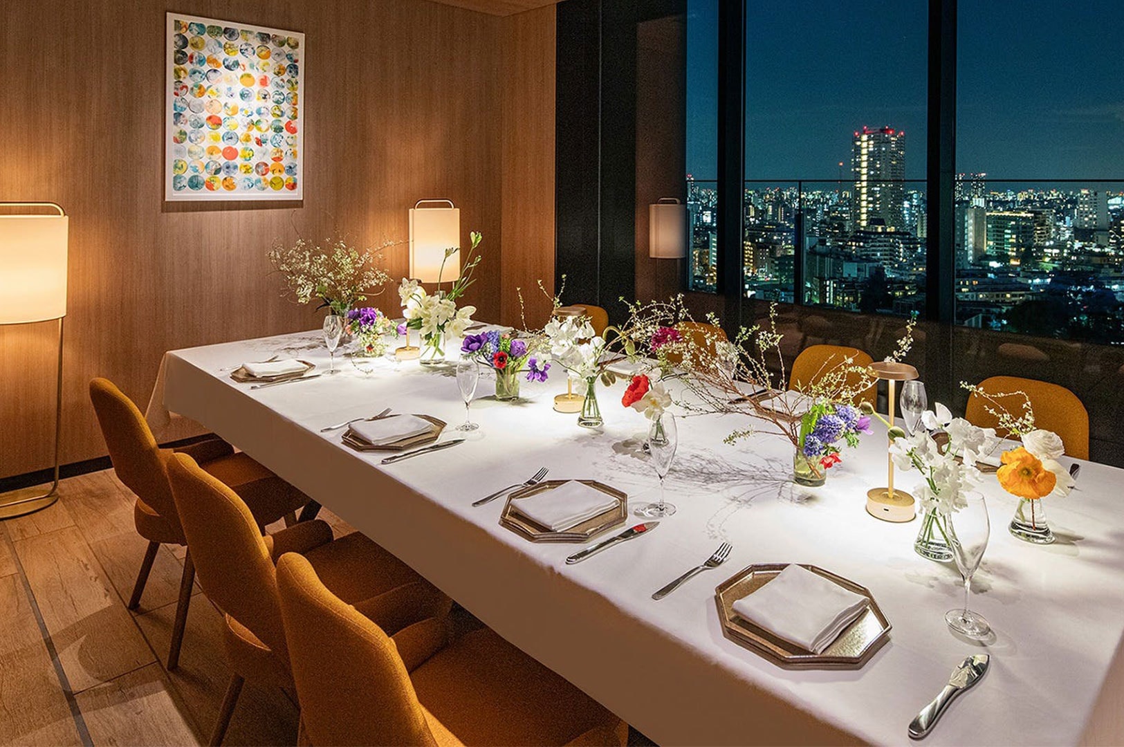A luxurious private dining table set with elaborate floral arrangements and ambient lighting, overlooking a sprawling city skyline at night.
