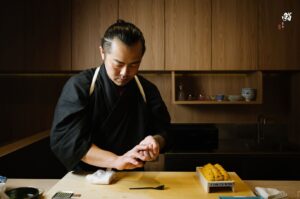 A focused sushi chef in traditional attire shaping rice for nigiri, with a wooden tray of fresh sea urchin (uni) on the counter.