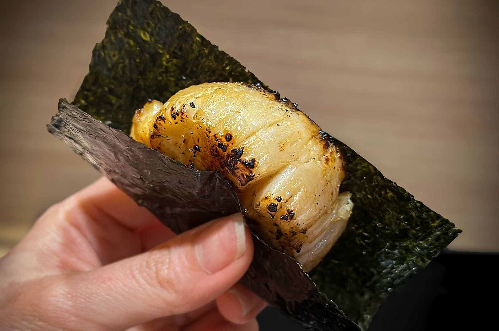 A hand holding a premium piece of sushi featuring a large, seared scallop (hotate) wrapped in crisp nori seaweed.
