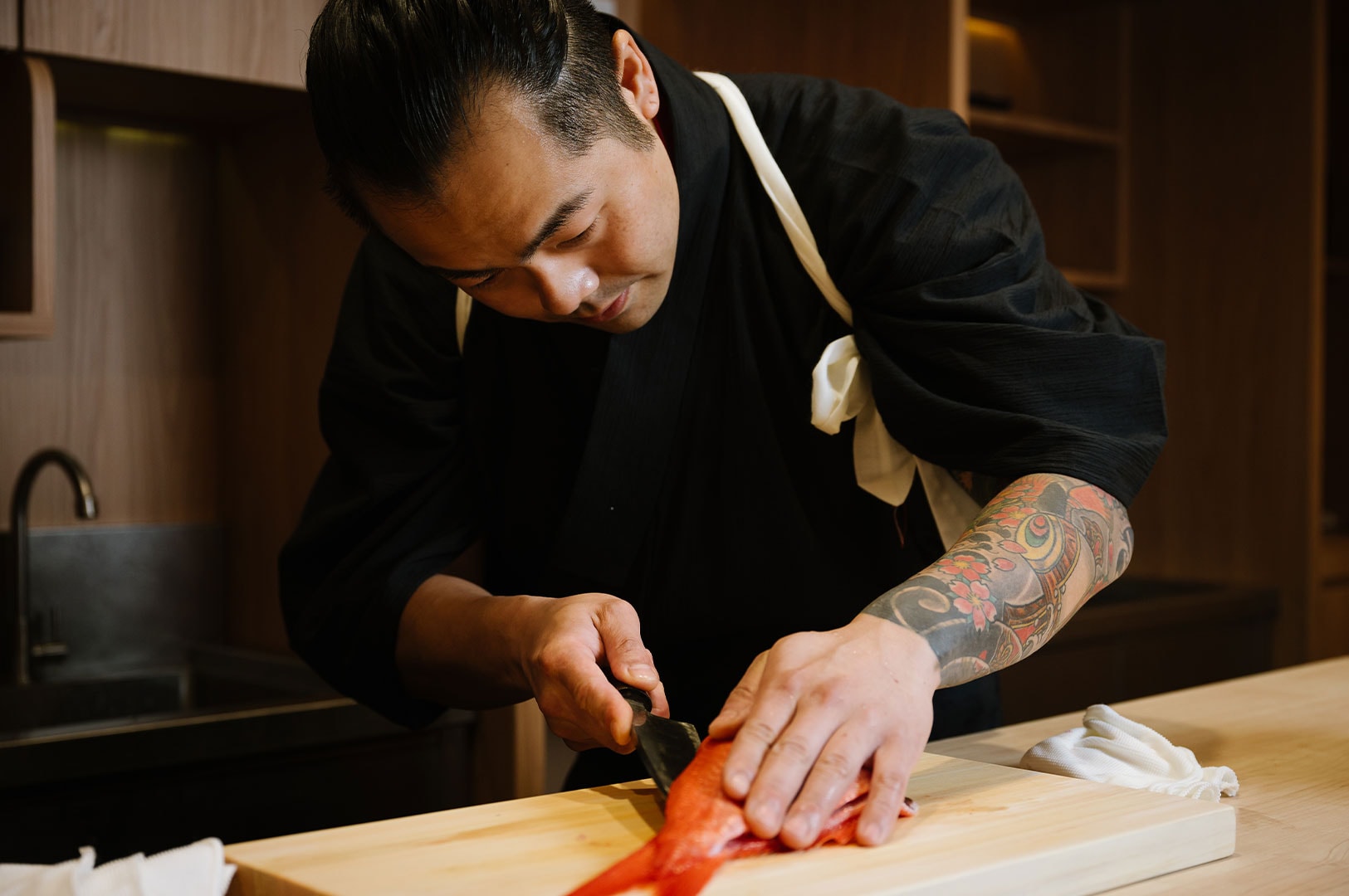 A focused sushi chef in a black kimono slicing a fresh red fish on a wooden cutting board using a traditional Japanese knife.