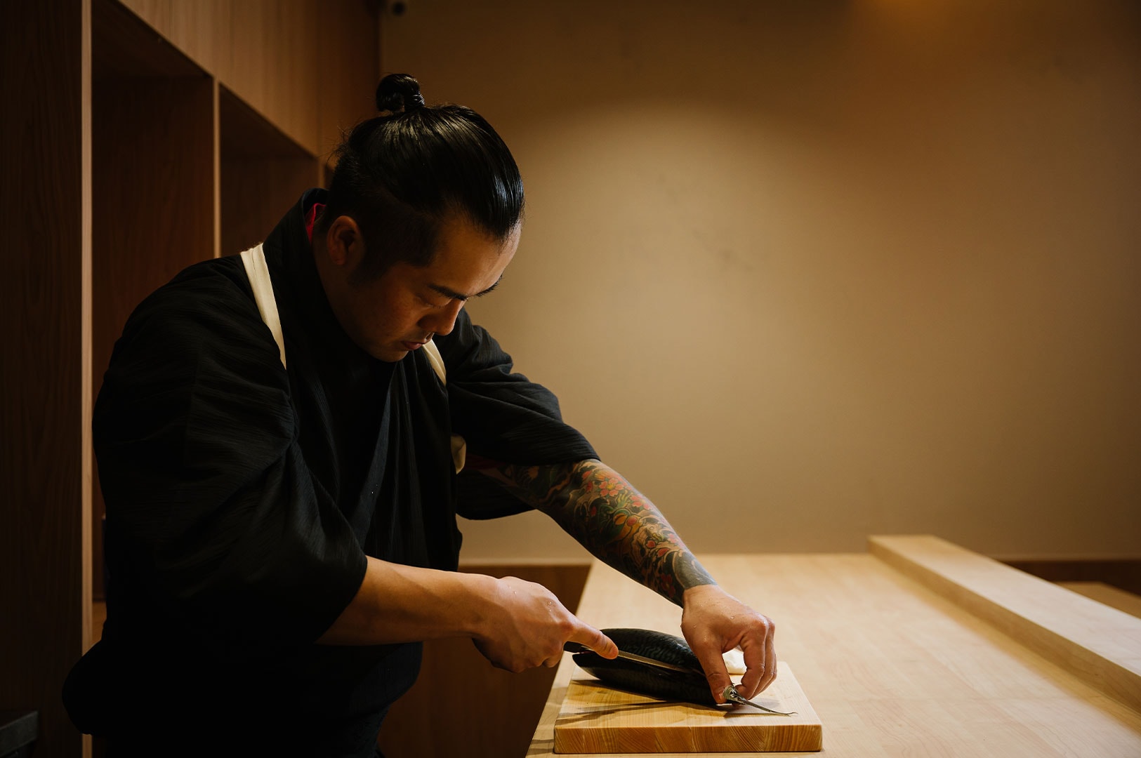 A traditional Japanese sushi chef with arm tattoos and a top knot focusing intensely while slicing a fish fillet on a wooden cutting board.