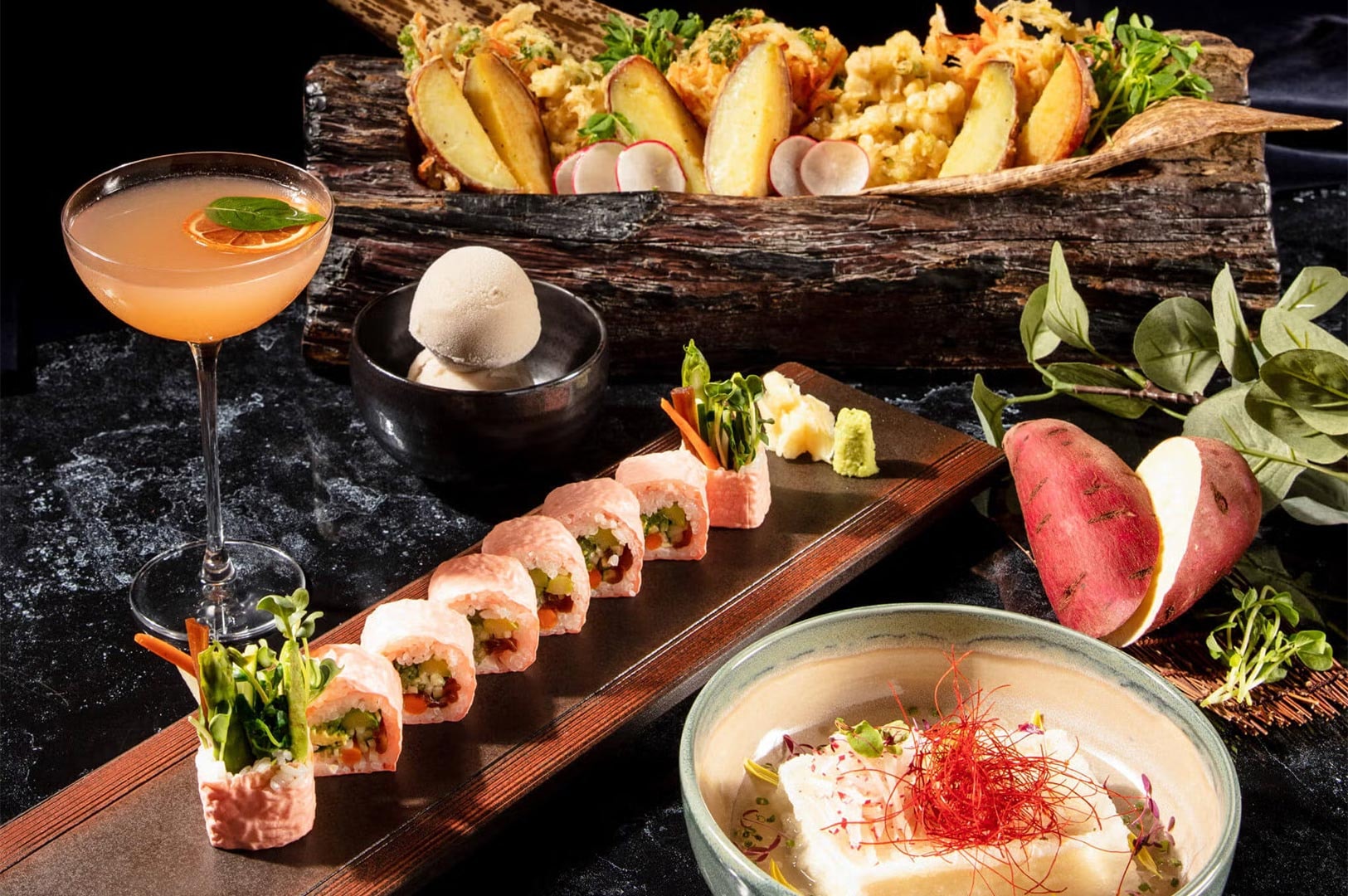 A gourmet Asian dining spread featuring pink soy-paper sushi rolls, crispy vegetable tempura, a craft cocktail, and dessert presented on rustic wooden serving boards.