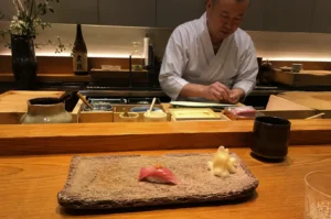 A skilled sushi chef in a white traditional uniform meticulously preparing nigiri behind a light-colored wooden counter in a dimly lit, minimalist Japanese restaurant. In the foreground, a single piece of tuna nigiri topped with a small garnish sits on a rustic, rectangular stone plate alongside a mound of pickled ginger, positioned directly in front of the viewer's seat. The background reveals the organized workspace of the chef, featuring various small wooden containers for ingredients, a bottle of sake, and soft, warm backlighting that highlights the clean lines of the wooden interior, creating an intimate and focused atmosphere typical of an omakase dining experience.