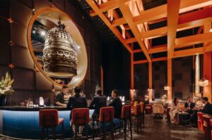 Interior of a high-end restaurant featuring a large circular mirror reflecting a giant bronze bell sculpture, distinctive orange geometric ceiling beams, and guests seated at a blue-lit bar.