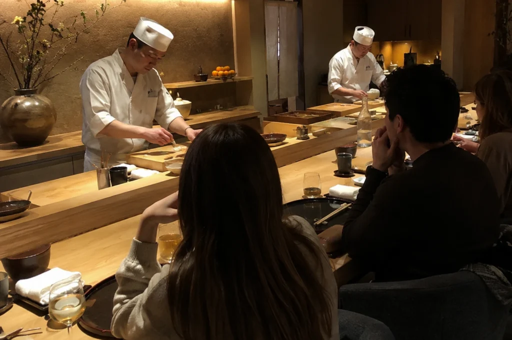 A wide shot of an intimate, high-end Japanese sushi counter with a warm, minimalist aesthetic. In the foreground, the backs of three diners are visible: a woman with long brown hair, a man in a dark sweater, and another diner partially out of frame. They are seated at a light-colored wooden bar. Across the counter, two sushi chefs dressed in traditional white uniforms and tall chef hats (tokin) are meticulously preparing food. The chef on the left is focused on a wooden cutting board, while the chef in the background is working near a service area. The background features textured, earth-toned walls, a large ceramic vase with budding branches, and shelves holding bowls of citrus fruit. The lighting is soft and golden, creating a serene, upscale atmosphere.