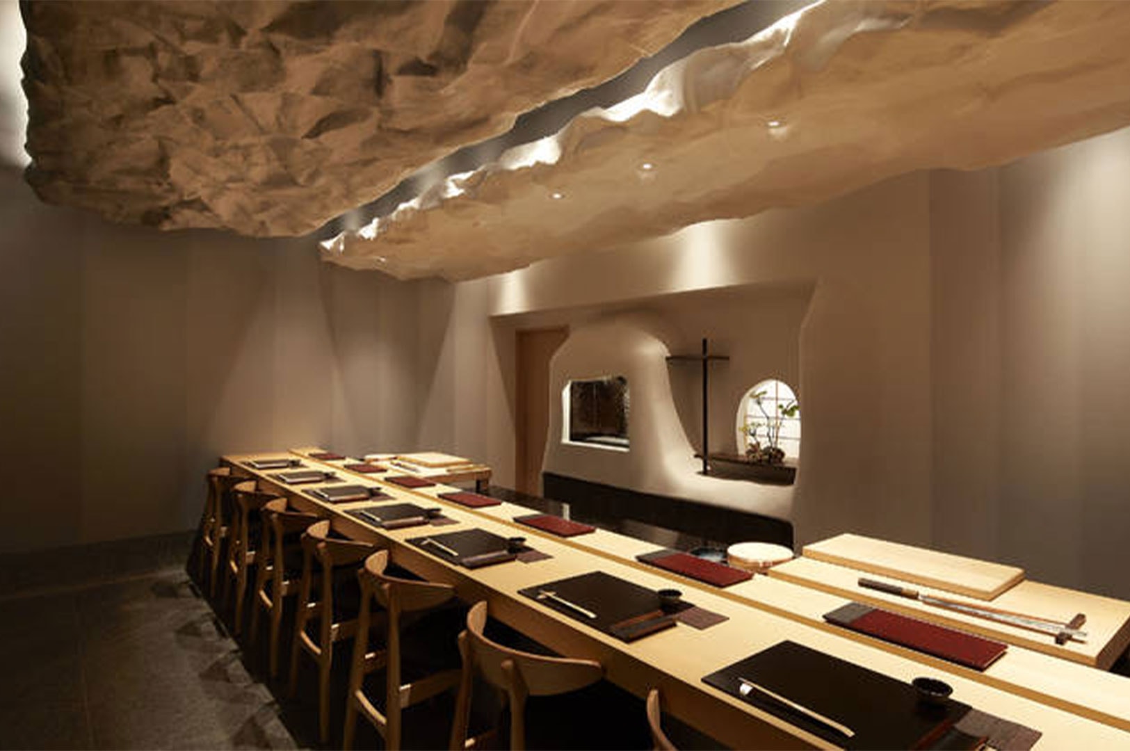 A minimalist sushi dining room featuring a long wooden table and a unique, white cloud-like textured ceiling installation.