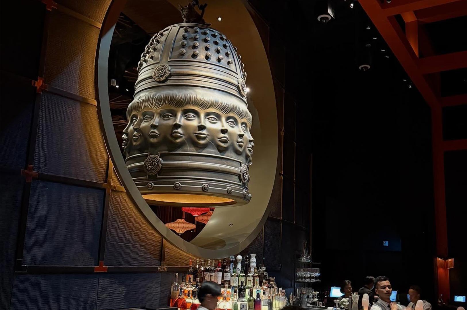 A massive, ornate bronze bell sculpture featuring multiple sculpted faces hanging prominently above a well-stocked bar in a luxury restaurant lounge.