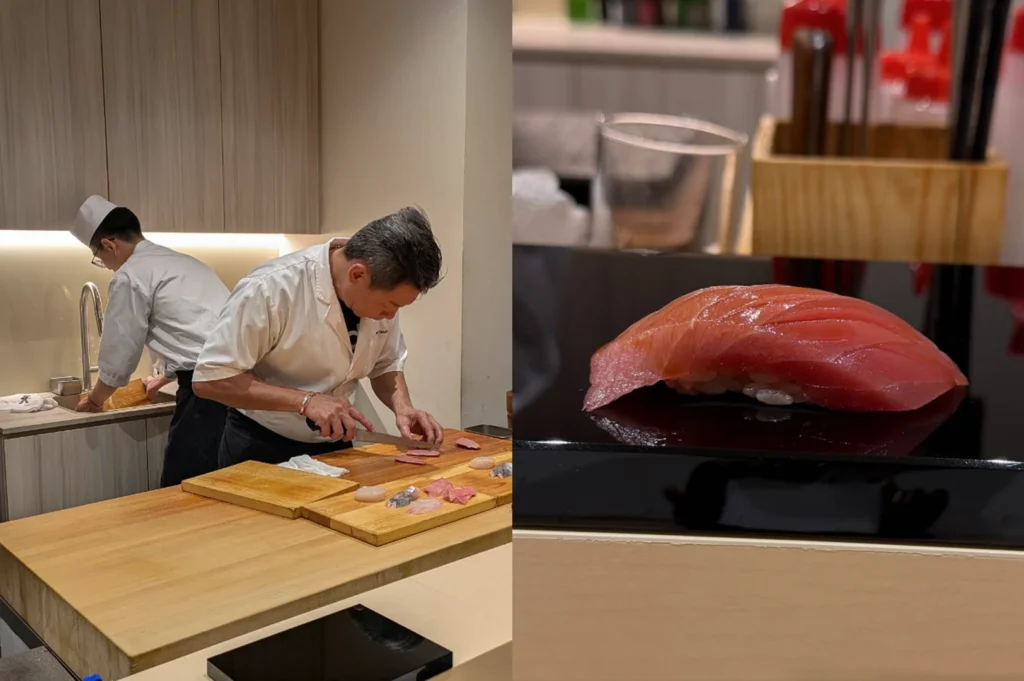 This split-screen composition focuses on the precision of sushi preparation and the final result. The left side shows a chef in a white short-sleeved coat leaning over a large wooden cutting board, expertly slicing translucent pieces of fish with a long, thin knife while an assistant works at a sink in the background. The right side features a dramatic, low-angle close-up of a single piece of otoro (fatty tuna) nigiri resting on a polished, reflective black lacquer stand. The fish has a deep pink hue with visible marbling and a glossy glaze, making it the centerpiece against a blurred background of kitchen supplies and glassware.
