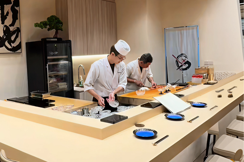 A minimalist Japanese omakase counter with light wood surfaces, warm recessed lighting, and five elegant grey chairs.