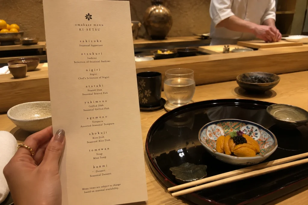 A close-up, first-person perspective at a sushi counter. A person’s hand, wearing a gold ring, holds a tall, cream-colored "Omakase Menu" titled "KI-SETSU." The menu lists courses including Sakizuke, Otsukuri, Nigiri, Atataki, Yakimono, Agemono, Shokuji, Tomewan, and Kanmi. In the lower right, a black lacquer tray holds a small, decorative ceramic bowl filled with bright orange uni (sea urchin) topped with black caviar and a small green garnish. Beside the tray are light wood chopsticks and a small leaf-shaped rest. The background is softly blurred, showing a sushi chef’s hands working on a wooden board and various tea cups and glassware on the wooden counter under warm ambient lighting.