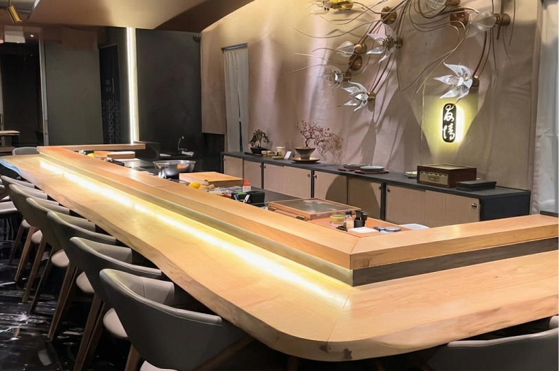 Two sushi chefs in traditional white uniforms preparing dishes behind a bright, modern L-shaped wooden counter.