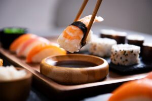 Close-up of chopsticks dipping a piece of shrimp nigiri into a wooden soy sauce bowl, with a sushi platter in the background.