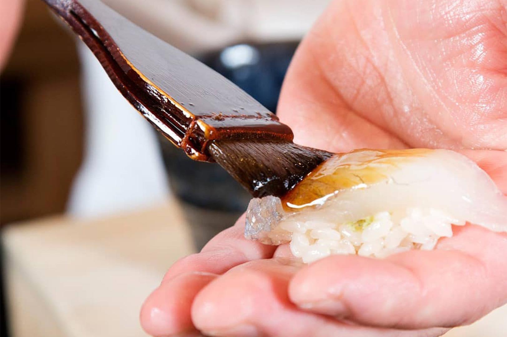 Close-up of a chef’s hand holding a piece of white fish nigiri while applying a dark nikiri soy sauce glaze with a traditional pastry brush.