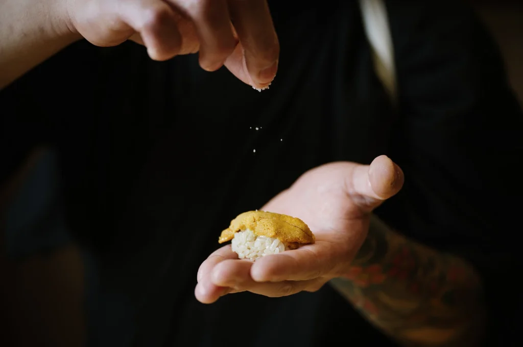 A sushi chef's hands carefully sprinkling seasoning onto a piece of fresh uni (sea urchin) nigiri.