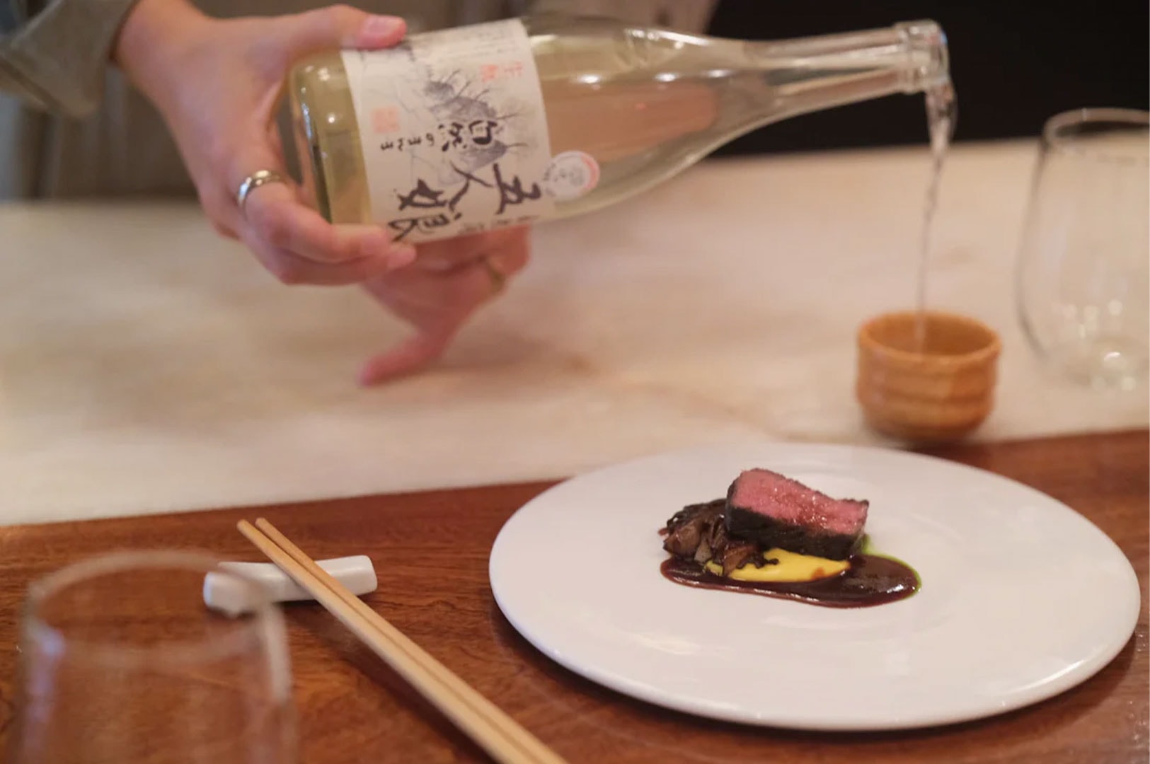 A server pouring premium sake from a bottle into a cup, accompanied by a plated gourmet dish in a fine dining setting.