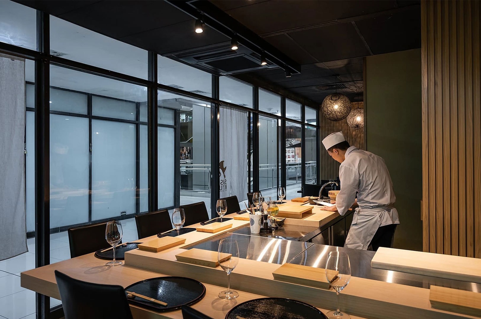 A sushi chef preparing food behind a clean wooden counter in a modern restaurant with floor-to-ceiling glass windows.
