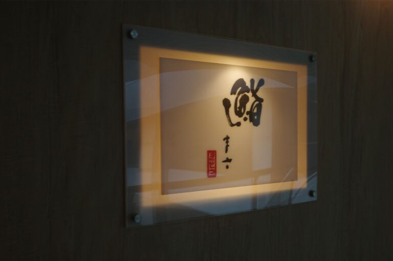 An illuminated, framed sign featuring traditional Japanese calligraphy logo on a wooden wall at the entrance of a sushi restaurant.